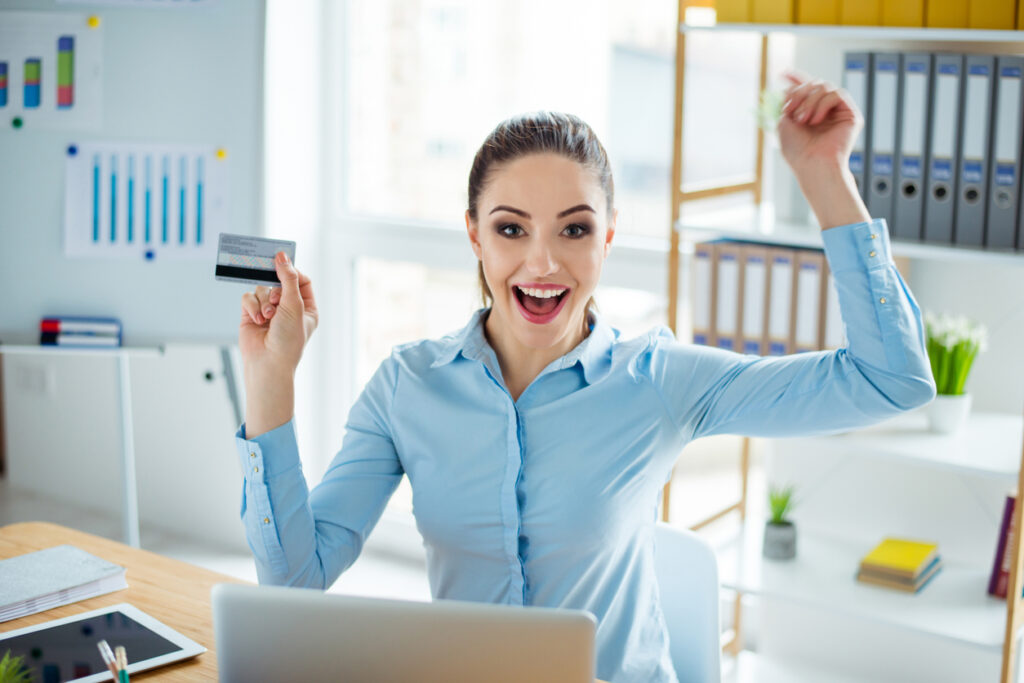 Mujer joven con tarjeta de crédito y triunfante con las manos levantadas mientras lograba acumular millas. Enfoque sus compras online y físicas a crédito para acumular puntos.