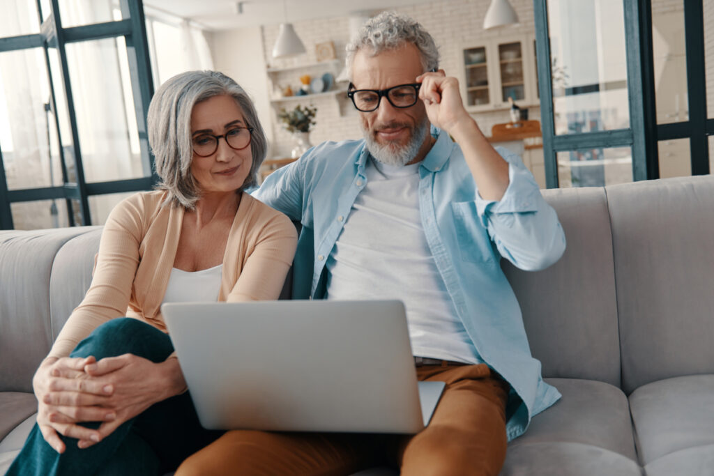 Una pareja de ancianos moderna con ropa informal sonriendo y usando una laptop mientras se une en casa. MercoMillas, la empresa que revolucionó el mercado de las millas.
