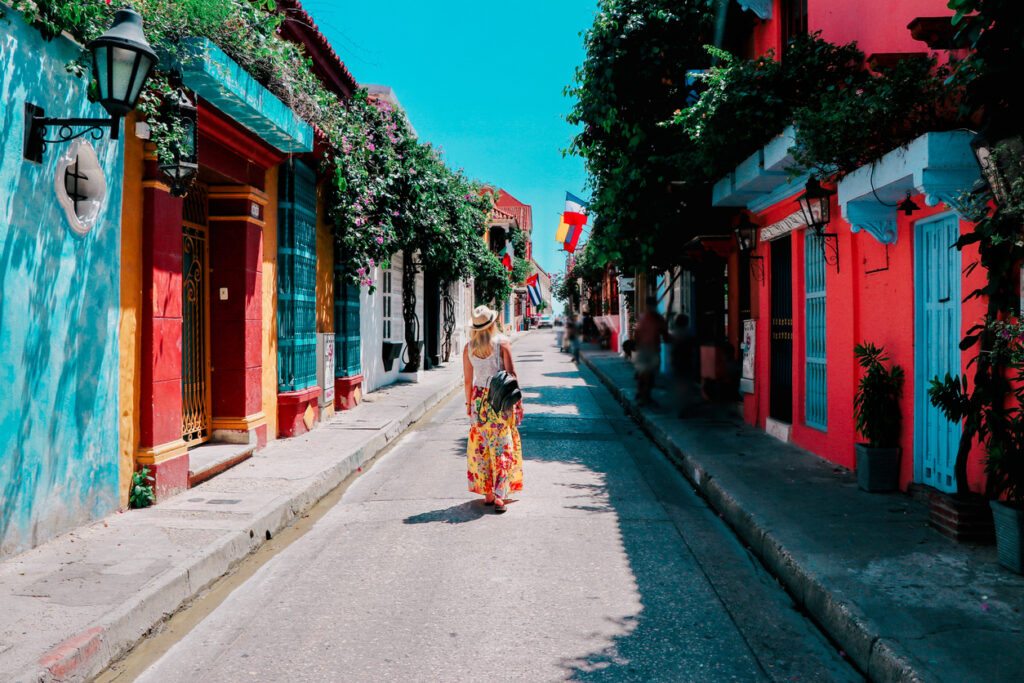 una mujer con blusa blanca, falda colorida, sombrero y mochila negra camina por una calle de Cartagena. Ella está haciendo un recorrido por los mejores destinos de América del Sur