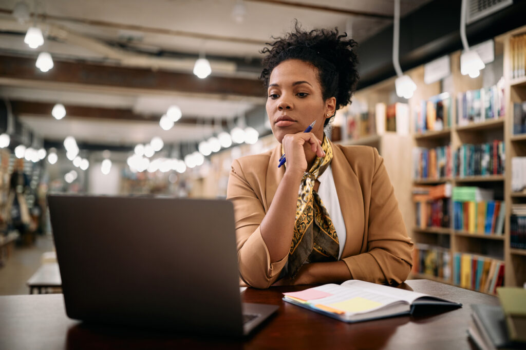 una mujer negra con el cabello recogido hacia atrás, viste una blusa blanca, un blazer marrón y una bufanda amarilla. Está en una biblioteca mirando su cuaderno y aprendiendo cuáles son los mitos sobre millas aéreas.