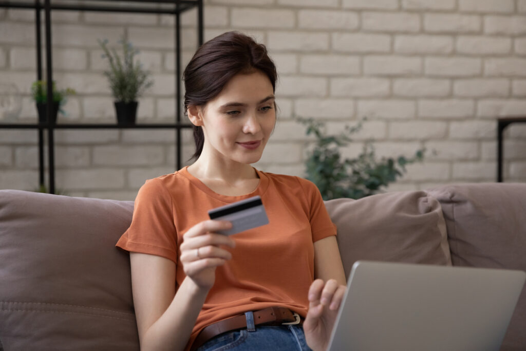 una mujer blanca, con cabello castaño recogido hacia atrás, ojos rasgados y una blusa naranja está sentada en el sofá, jugando con su cuaderno mientras sostiene una tarjeta. Ella está revisando la nueva regulación de millas con tarjeta BBVA