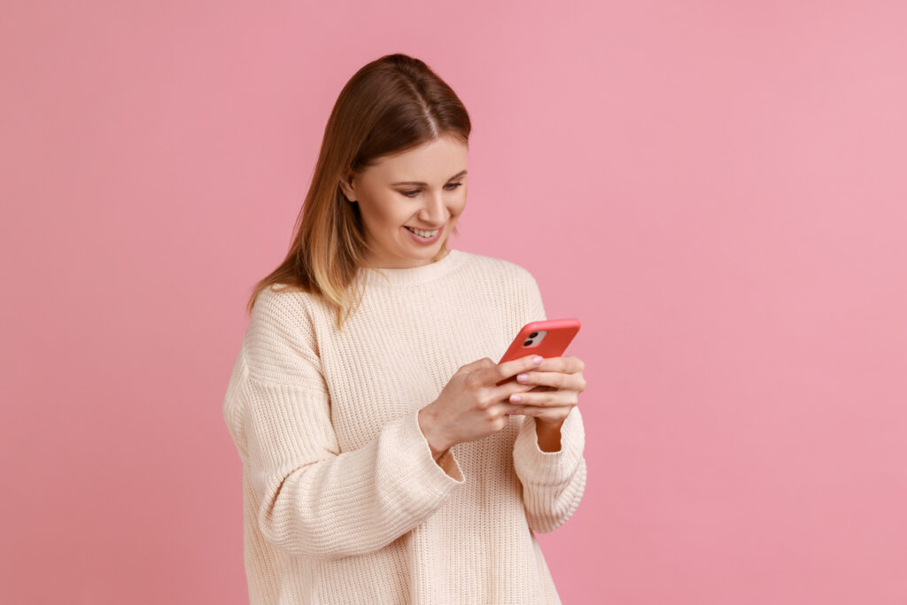 Fondo rosa, una mujer blanca y rubia con una blusa de punto blanca sostiene un teléfono celular mientras sonríe mirando la pantalla. Está feliz porque descubrió las ventajas de la venta de millas aéreas