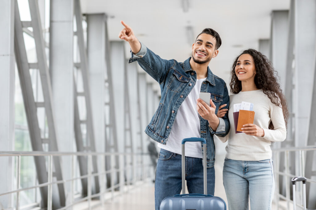 Una pareja, formada por una mujer blanca, con cabello largo y rizado y café oscuro, camisa blanca con mangas largas y jeans claros y un hombre blanco, con cabello castaño oscuro corto, barba, camisa blanca, chaqueta de mezclilla y jeans está de pie en una salida salón. La mujer sostiene dos pasaportes mientras que el hombre señala hacia un lado. Los dos están felices porque lograron viajar después de que aprendieron a acumular millas aéreas.